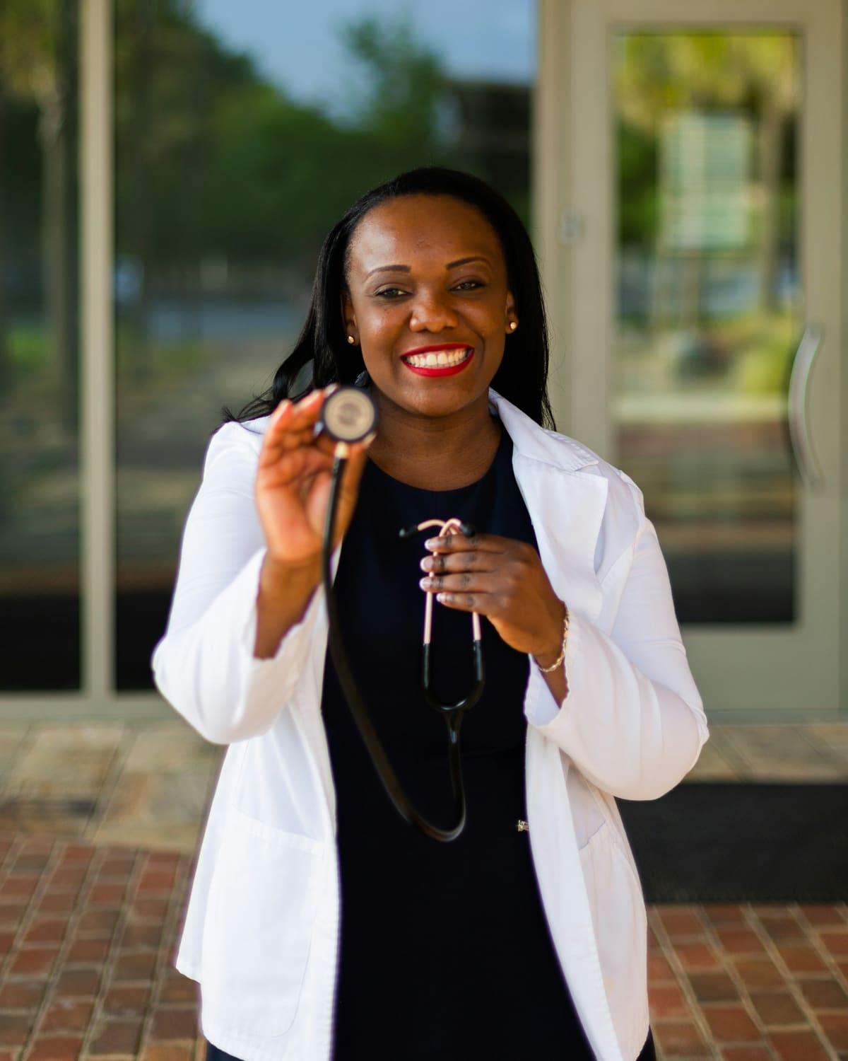 A doctor in a white coat smiles and holds up her stethoscope.