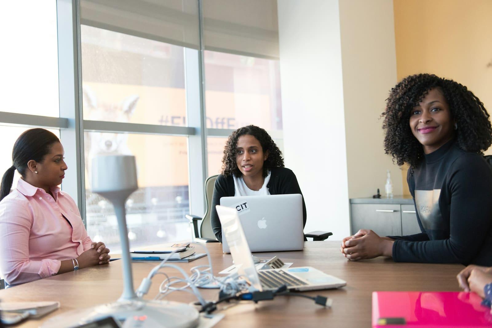Three women in a meeting at an office table with a laptop.